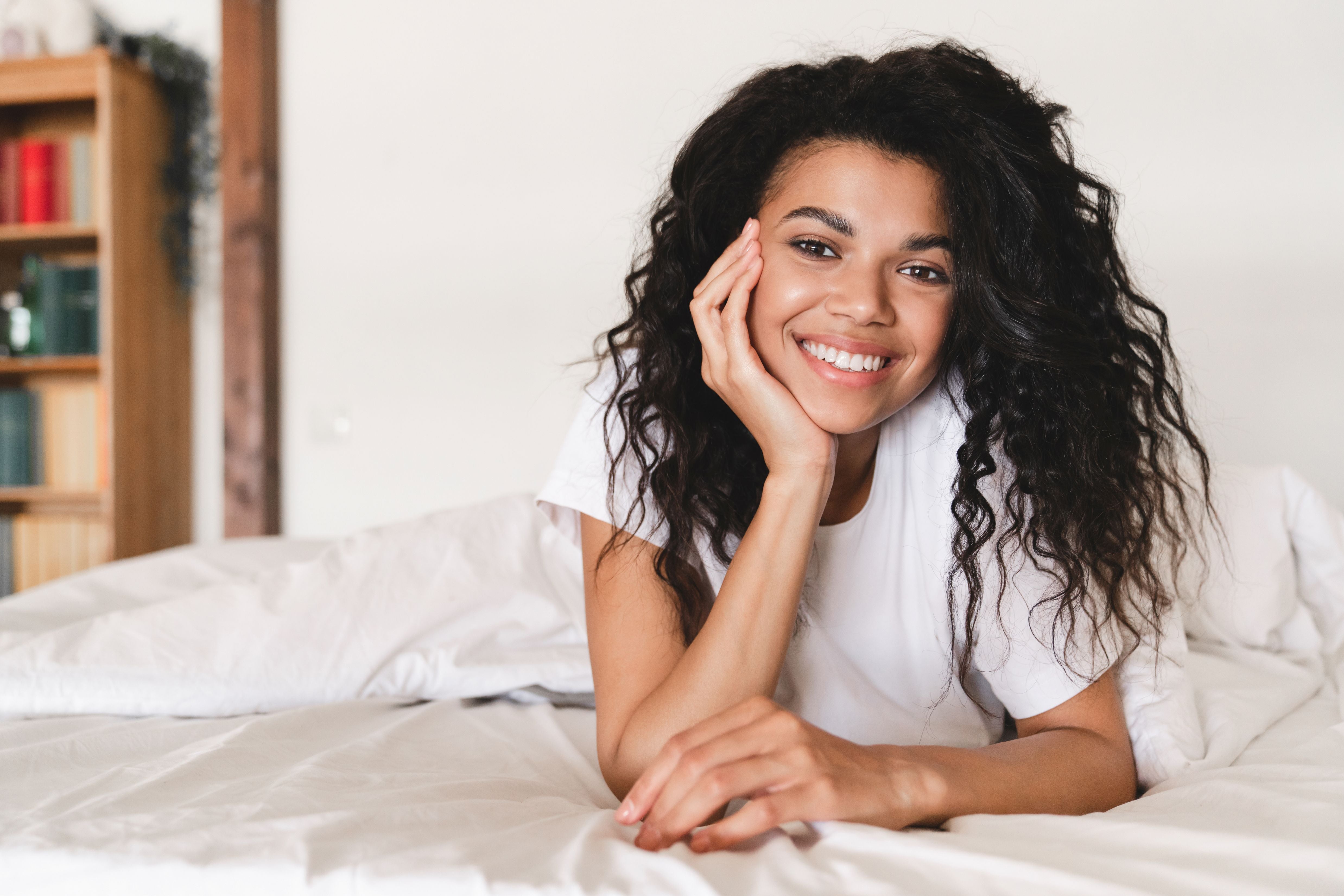 Woman lying on a bed with a smile, wearing a white t-shirt.