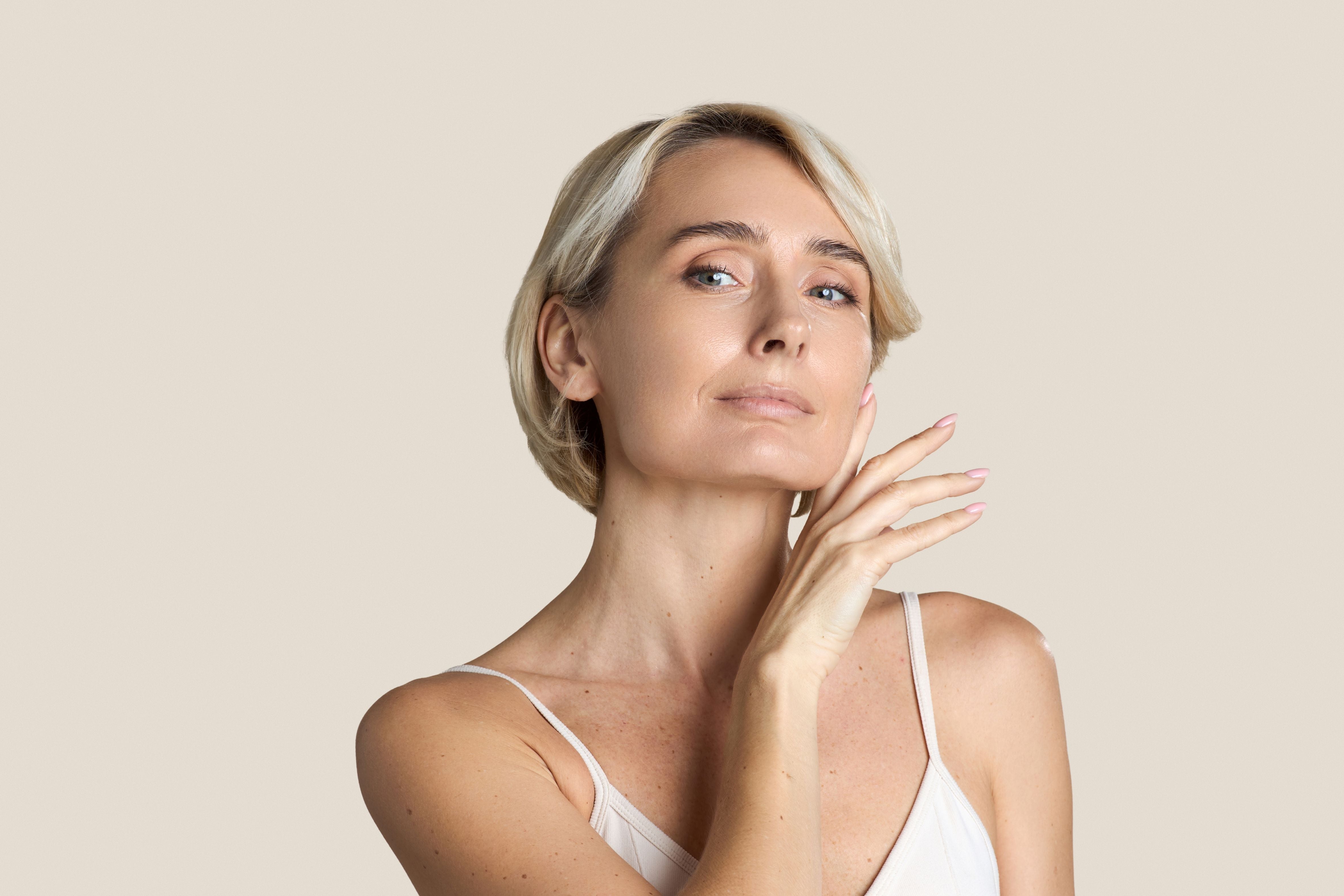 Woman with short blonde hair wearing a white tank top against a beige background